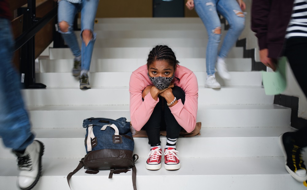 2021 national school climate survey: young black student with facemask sitting on steps sadly while other students walk by