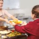 health school meals and food grants: lunch service woman hands plate of food to girl in red jacket