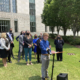 Transgender youth treatment: Crowd of people in business casual wear stand on a green lawn bedind a young asult with blonde hairwearinga blue shirt and tan pants, speaking into a microphone to a crowd off-camera.