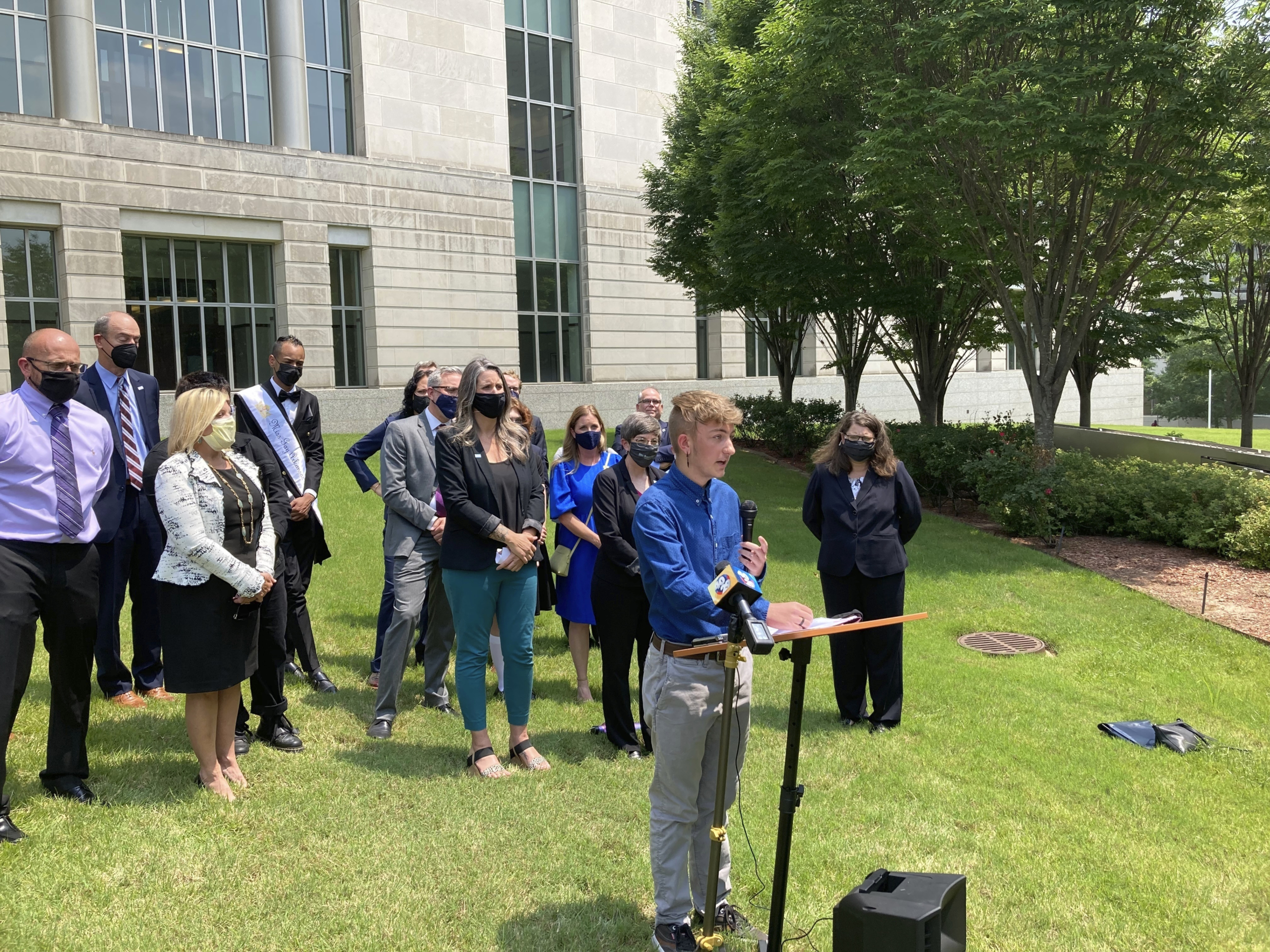 Transgender youth treatment: Crowd of people in business casual wear stand on a green lawn bedind a young asult with blonde hairwearinga blue shirt and tan pants, speaking into a microphone to a crowd off-camera.