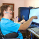 Disabled voters rights_Young, blonde, white man in blue t-shirt sits in wheelchair wearing headset and reaching to touch computer screen