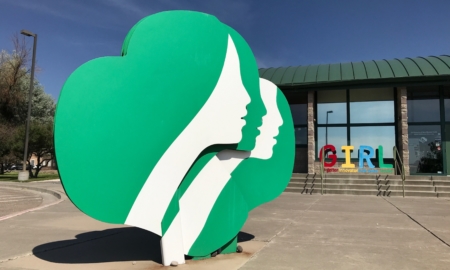 Girl Scout Donation: Large light green and white Girl Scout sculptural 3d logo sits on the ground outside a one-story wood building