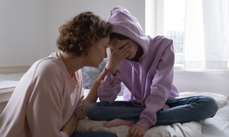 Child Anxiety Treatment: Asult woman with short brown hair crouches next to young teen chile sitting cross-legged in bed covering face with hand wearing lavendar hoodie with hood up and blue jeans
