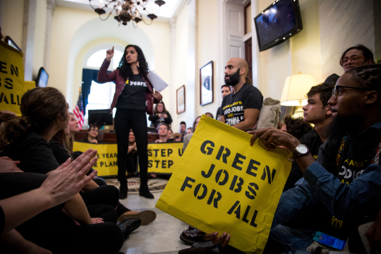 Youth climate activists: Young adults and teen sit on the floor in large white hallway holding yellow signs listening to a standing speaker