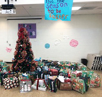Foster care community: A large decorated Christmas tree is surrounded by numerous wrapped presents below a sign that reads "Tis the season to be foster"