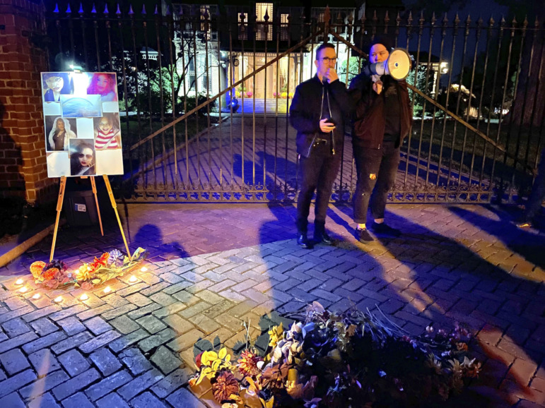 Transgender Remembrance: Two people in dark clothing stand speaking into megphone on brick sidewalk with wreath of lights and easel with photos of 5 people.