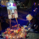 Yransgender rembrance: Oersin with white hair kneels on sidewalk next to wreath with loghts and easel with photos of 5 people