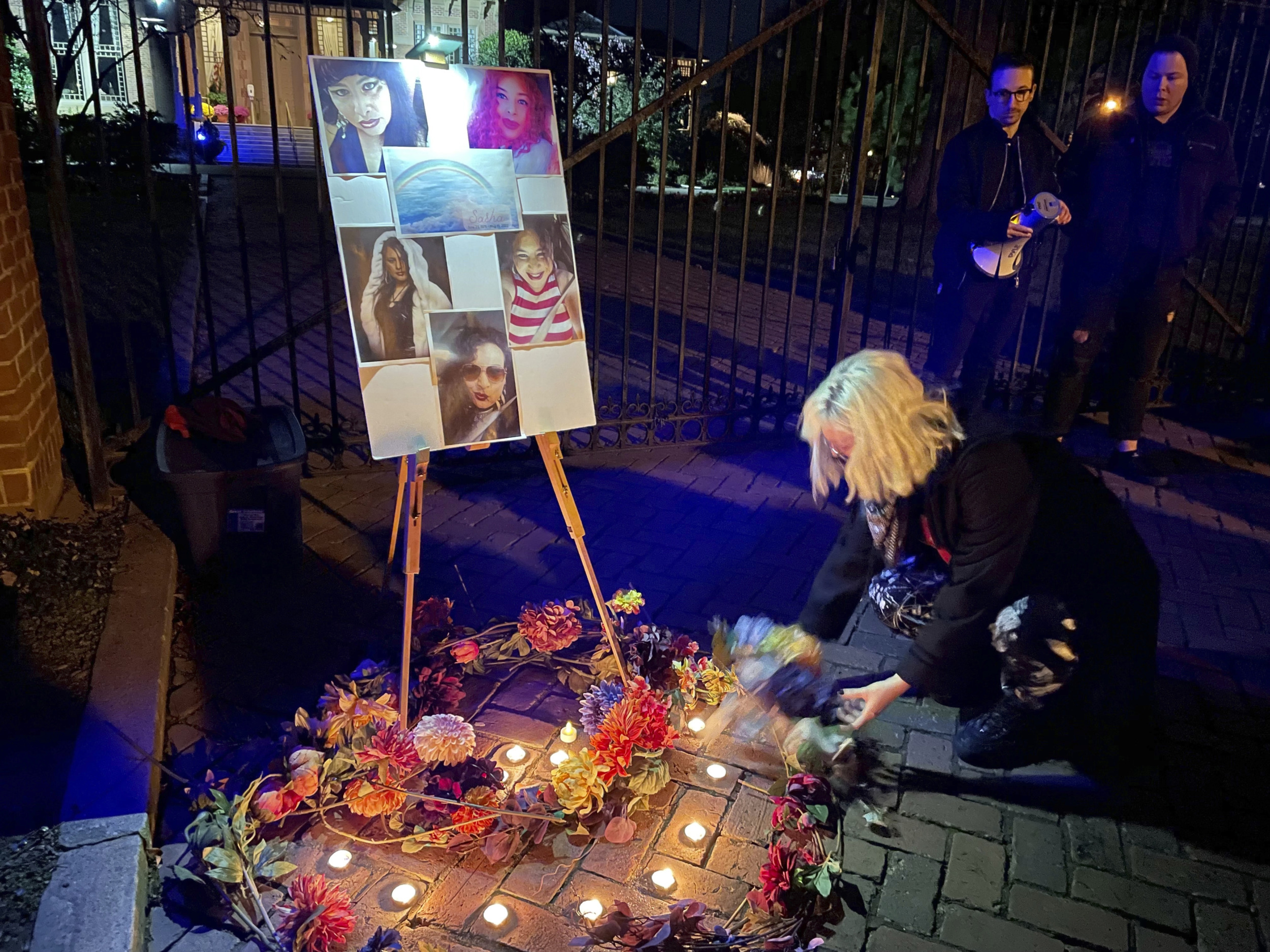 Yransgender rembrance: Oersin with white hair kneels on sidewalk next to wreath with loghts and easel with photos of 5 people