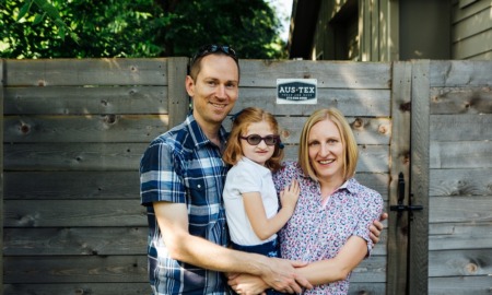 specialized dental care for disabled youth: parents smiling while holding child wit sunglasses in front of wooden fence