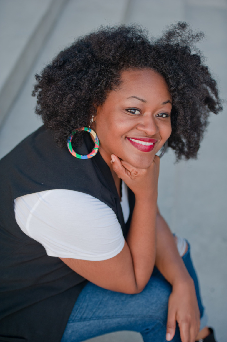 YTAC_Bianca Baldrige Headshot: Black woman with thick black hair in navy vest and white top has head resting on hand smiling into camera