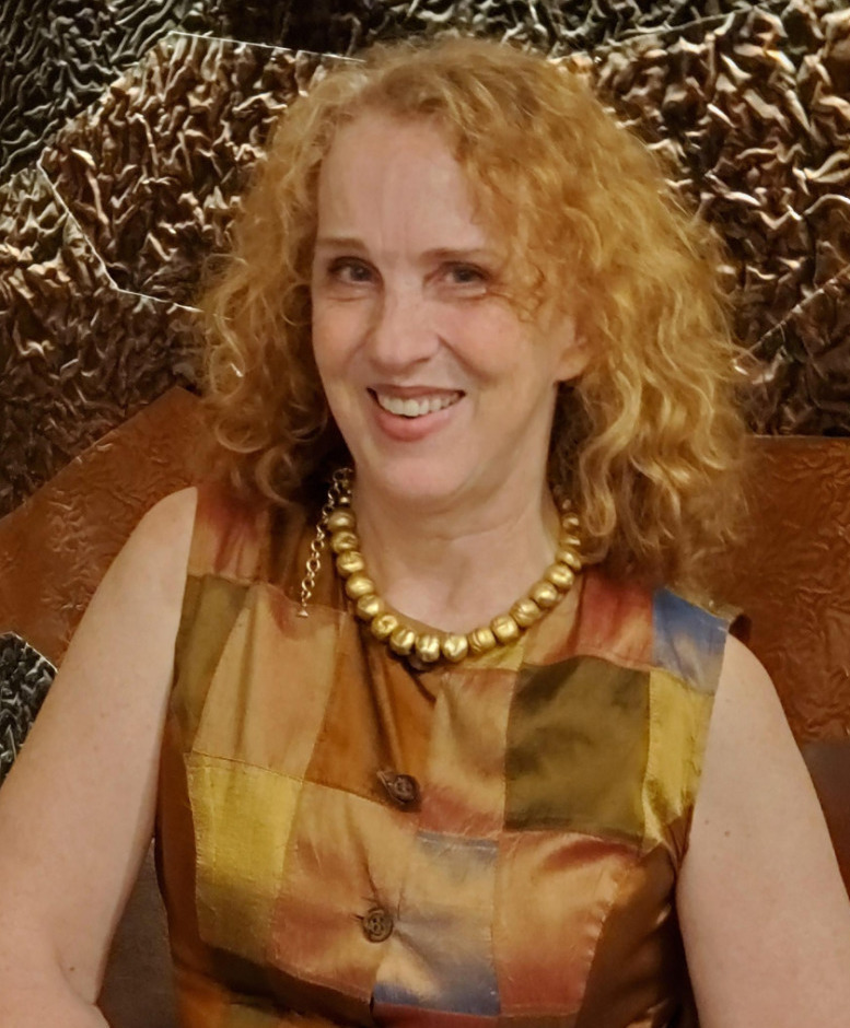 Merita Irby headshot: Wiman with blonde. curly hair wearing sleeveless gold print dress smiles into camera