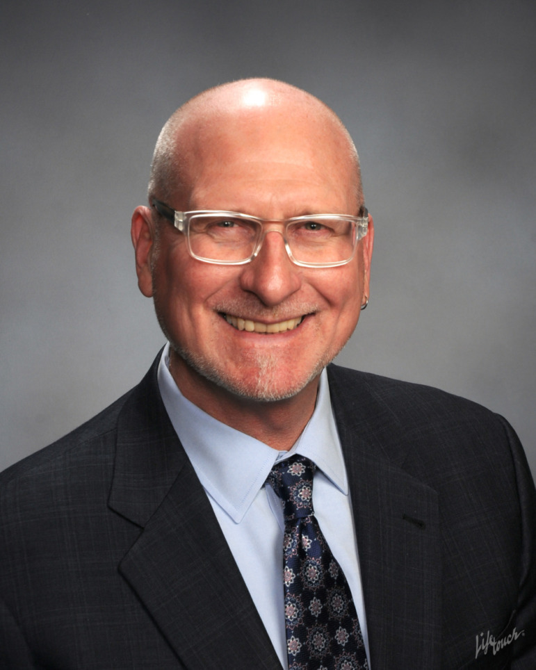 Michael Funk headshot: Older, balding man with wire-rim glasses in baxck suit and tie with light gray shirt smiles into camera