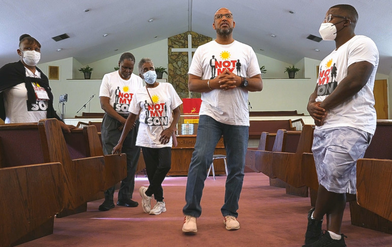 Gun violence curge: Black man and group of Black adults and teens wearing Not My Son t-shirts stand in a church aisle meeting