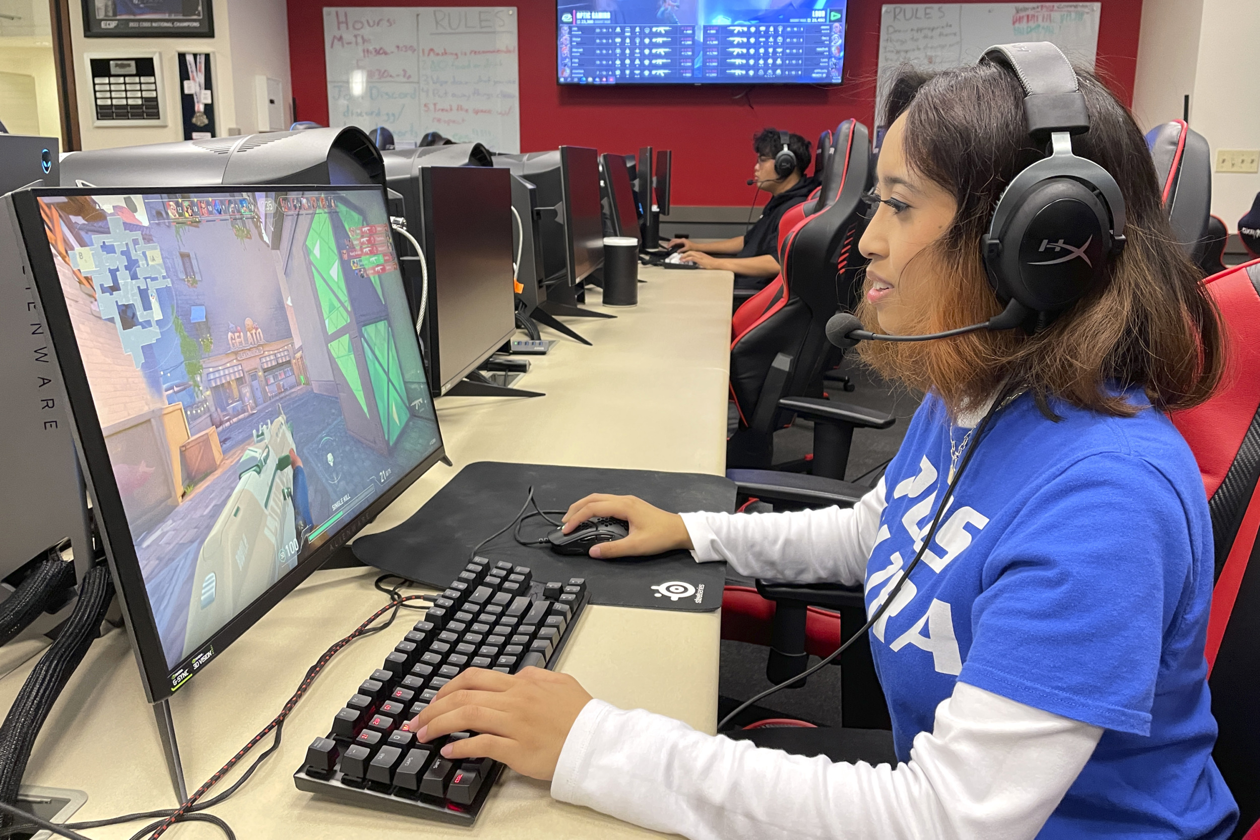 eSports: Young womans with dark hair wearing a headset and blue t-shirt sits at desk with large monitor and gaming keyboard playing game