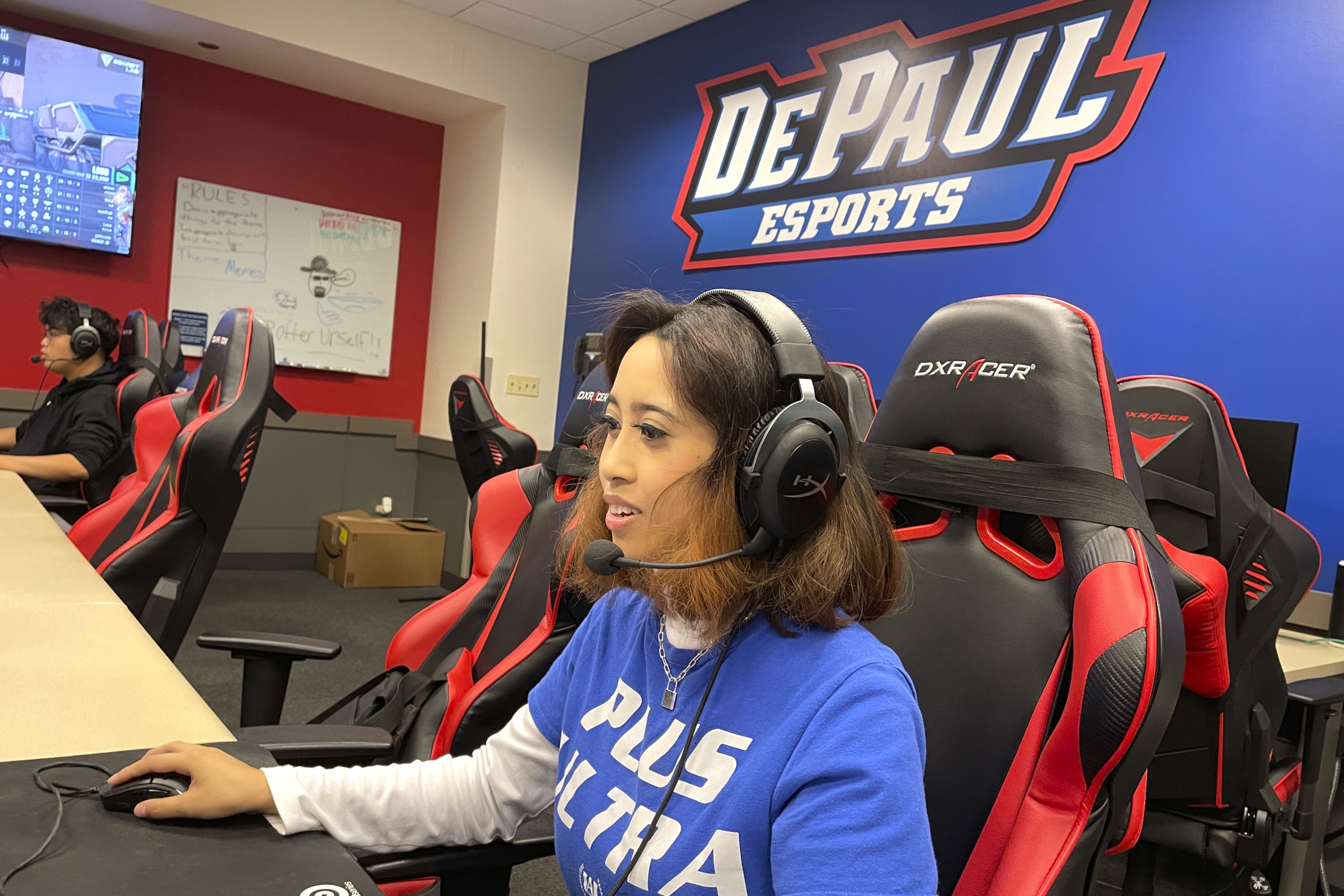 Esports: Young woman wearing headset sits at large computer screen in gaming room with multiple computers.