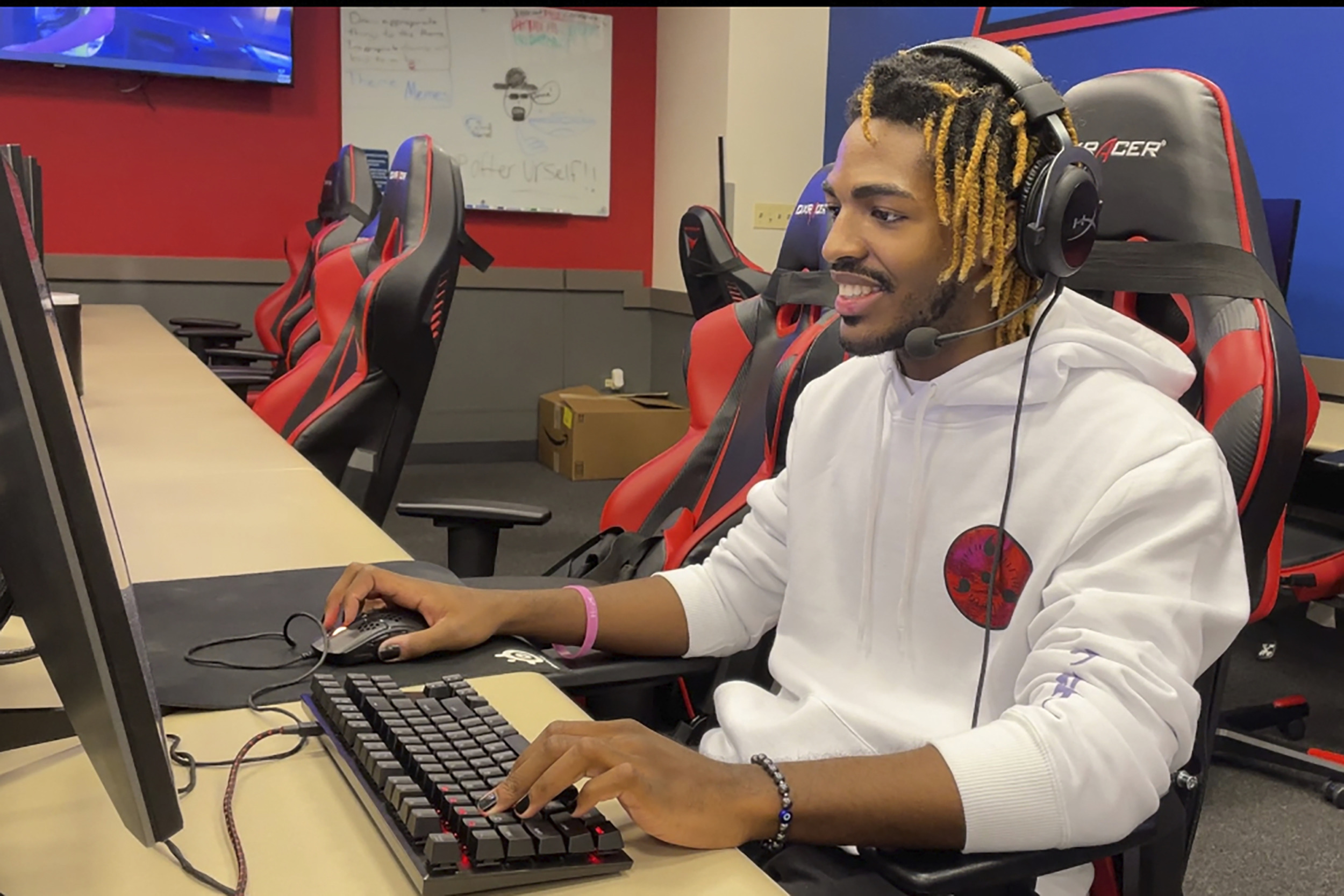 eSports: Young Black man with blonde dreads wearing headset and white sweatchirt hoodie sits ar desk with computer monitor using gaming keyboard in red and blue room with many computers