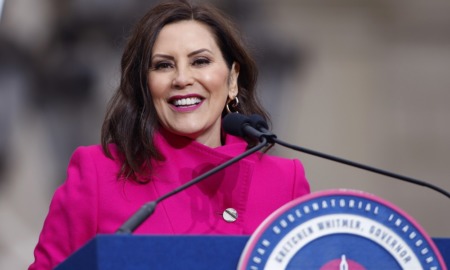 Michigan Gov. Whitmer calls for prek for all 4 year-olds: brunette woman in pink jacket smiling at podium with microphones