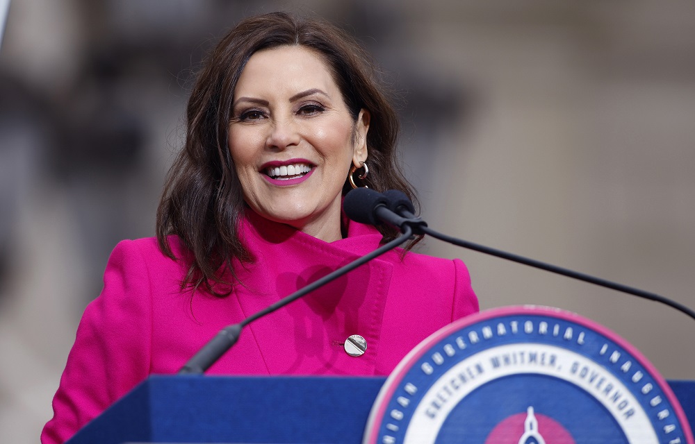 Michigan Gov. Whitmer calls for prek for all 4 year-olds: brunette woman in pink jacket smiling at podium with microphones