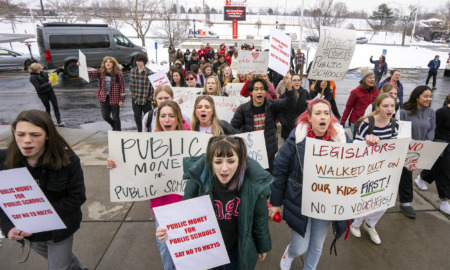 School Funding: Large group of young people in winter clothing stand on a sidewalk with sow in the background holding hand-made signs with language about school funding.