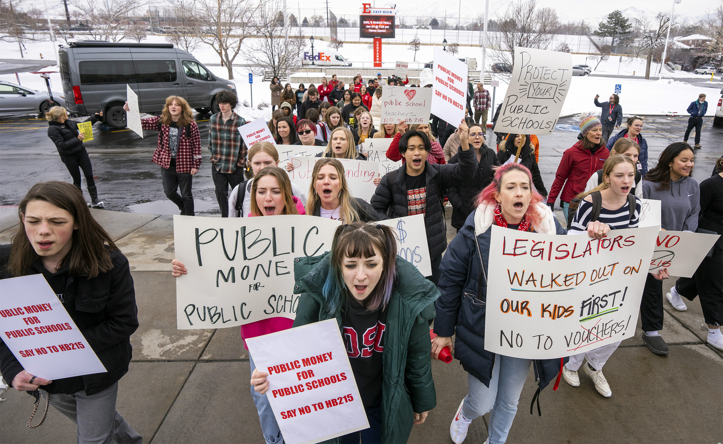 School Funding: Large group of young people in winter clothing stand on a sidewalk with sow in the background holding hand-made signs with language about school funding.