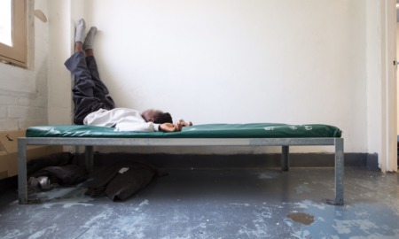 Juvenile detention populations low: Young black teen lies on bed with legs propped up on wall on cot in empty room with grey cement floor and white walls