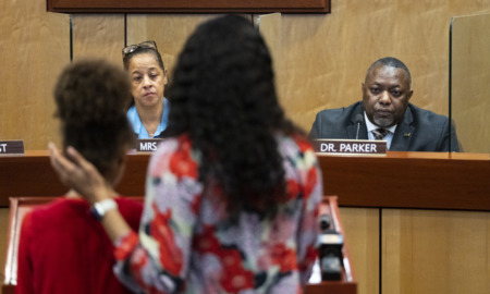 Student attacks teacher: White man and Blick man sit feacing camera behind long, light wood podium with two dark-haird woman standing facing them.