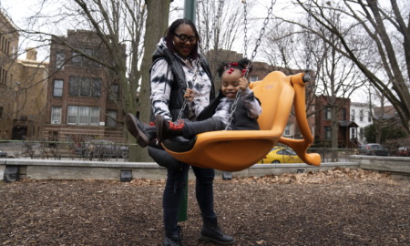 Childcare: Black woman with dark hair and black-framed glasses pushes toddler in bright yellow s playground swing