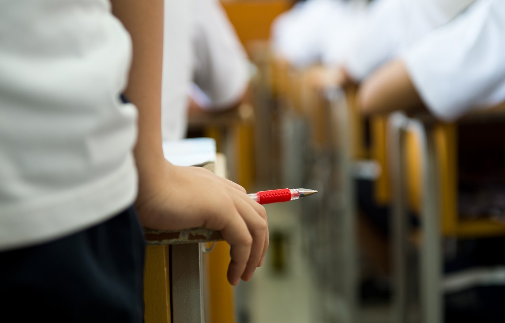 Schools face pressure to take harder line on discipline: closeup of students hand holding red pen with class in desks in background