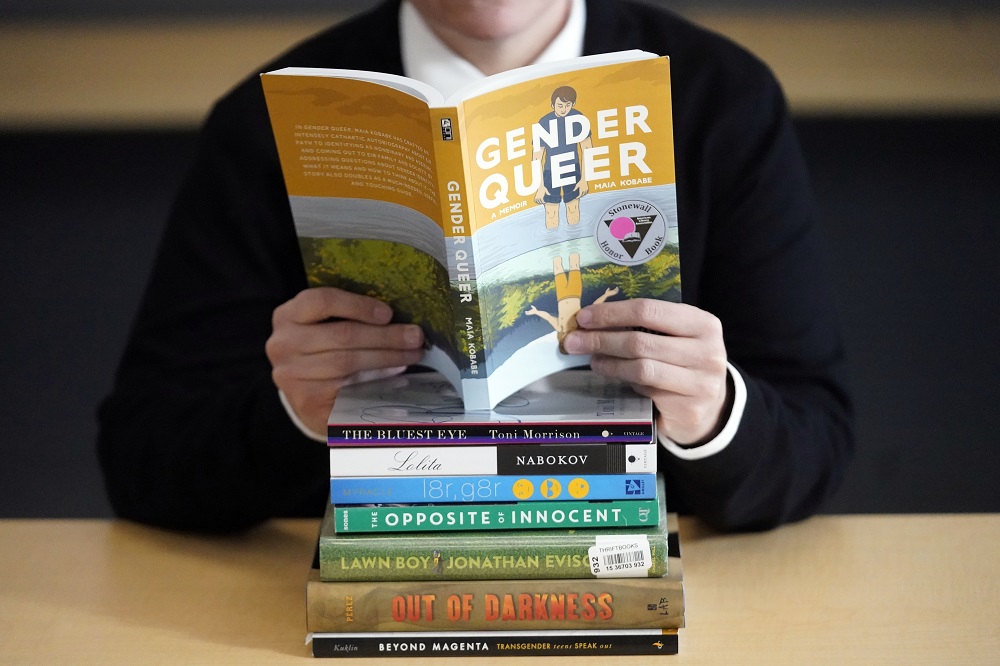 Walking on eggshells; teacher's responses to classroom limitations: person holding a book titled "Gender Queer" over a stack of other books