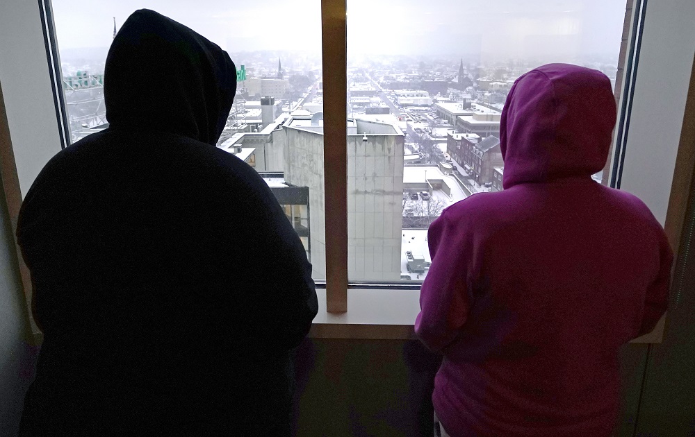 Ex-state adviser implicated in youth center abuse lawsuits: two hooded people look out at city from high window