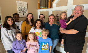 Foster care mental health: Family standing inside a house with nine children from early elementary to high school age stand closely togerher with a male and female adult.