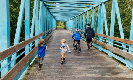 Telehealrh: Three young children and adiult wearing winter coats run away from camera across eooden bridge with terquoise framing on sides and overhead