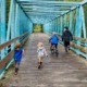 Telehealrh: Three young children and adiult wearing winter coats run away from camera across eooden bridge with terquoise framing on sides and overhead