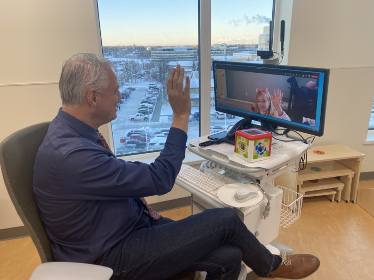 Telehealth: Older gray-haired man sits at desk waving at people on computer monitor in online meeting