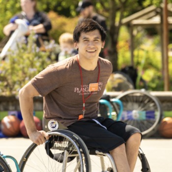 Asaptive sports: Teen with dark cury hair sits outdoors in wheelchair wearing black shorts and brown t-shirt
