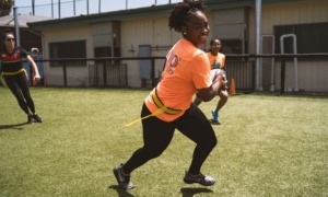 girls of color sports participation grants: black girl in orange shirt smiling while running with a football