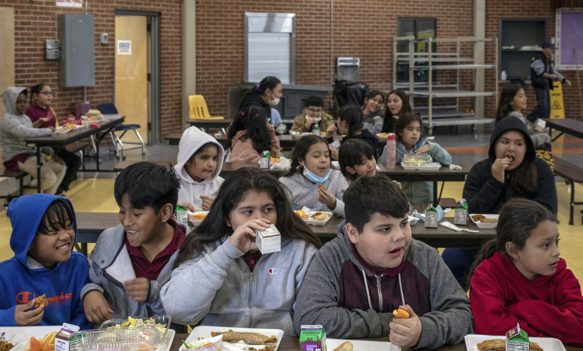 Hungry kids: Elementary school students sit at several lunch tables inside a school cafeteria with red brick walls.