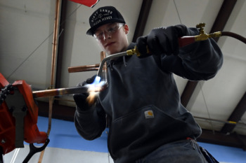 Skipping College: Young adilt in dark wirk uniform and baseball cap weldiing two copper pipes together