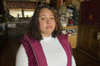 Skipping College: Young asult with long brown curly hair in deep pink winter vest and white turtlenecjk dtans in front of cluttered kitchen counter