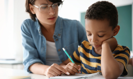 Tutoring: Young asult in clear-framed glasses and light blue shirt sits next to young child in gold/navy striped t-shirt as they review schoolwork on desk