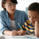 Tutoring: Young adult in clear-framed glasses and light blue shirt sits next to young child in gold/navy striped t-shirt as they review schoolwork on desk