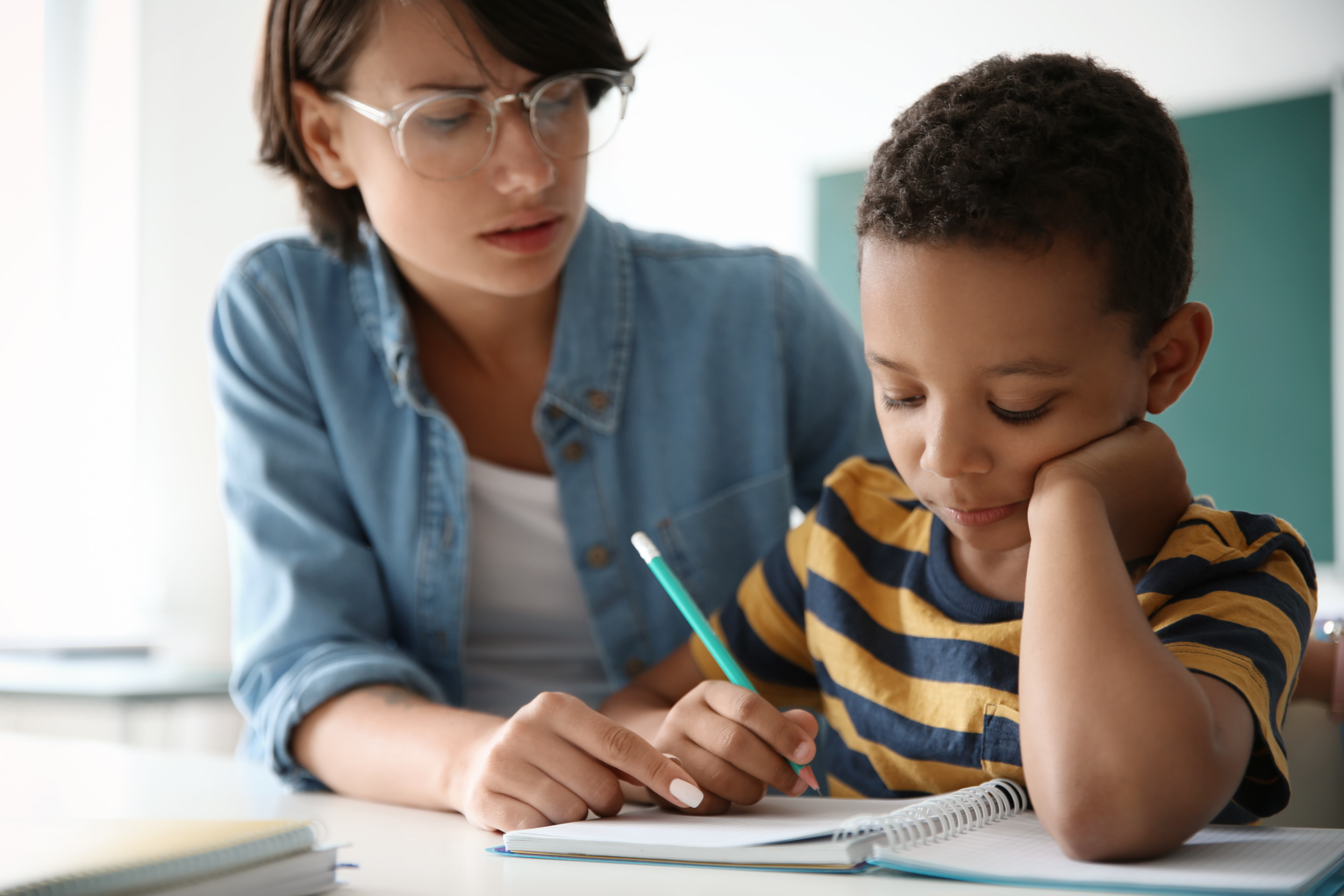 Tutoring: Young adult in clear-framed glasses and light blue shirt sits next to young child in gold/navy striped t-shirt as they review schoolwork on desk