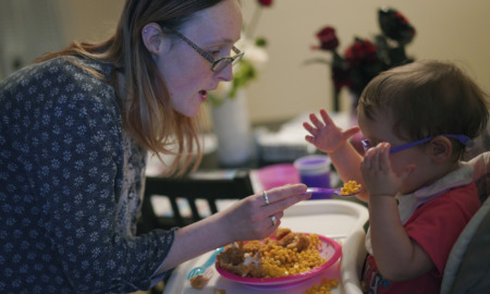 AI Flags Disabled Parents: Blonde woman with dark-framed glasses in print blouse holds spoon feeding infant in high chair from bowl of food.