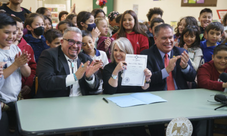 New Mexico: Large group of children stand behind two men and a woman, all in dark business suits sitting at mint green table, the men clapping and the woman holding up a paper in her hand