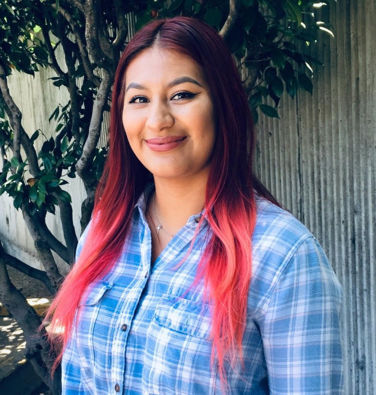 Gun Violence: Older teen with long auburn and pink hair in a blue and white plaid shirt stands outside by fence and tree smiling into camera