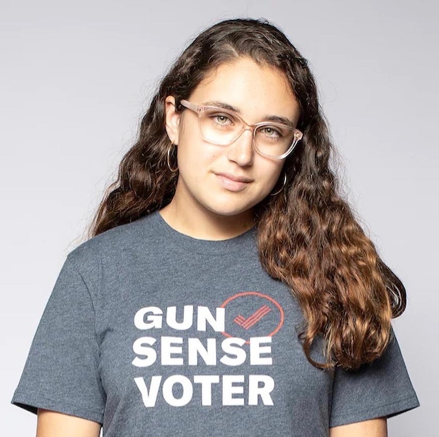 Gun Violence: Older teen with long brown hair and light-framed glasses in gray t-shirt with words "Gun Sense Voter" stands looking into camera
