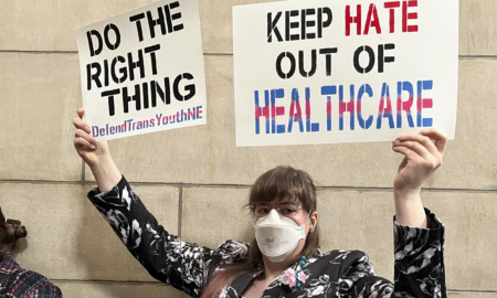 Transgender laws: Person in facemask with brown hair and glasses wearing black and white print jacket holds up two large signs "Do the right thing" and "Keep hate out of healthcare"