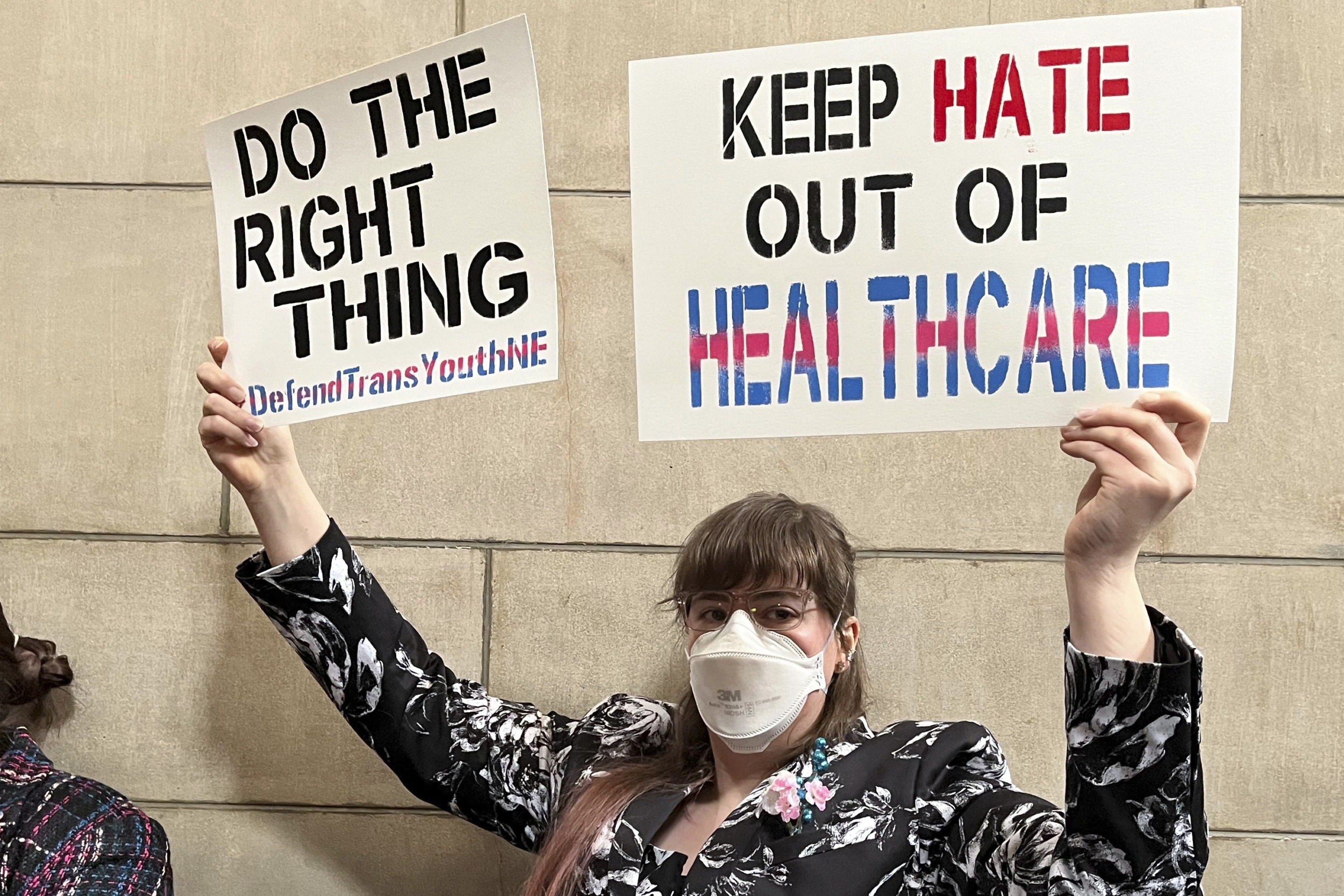 Transgender laws: Person in facemask with brown hair and glasses wearing black and white print jacket holds up two large signs "Do the right thing" and "Keep hate out of healthcare"