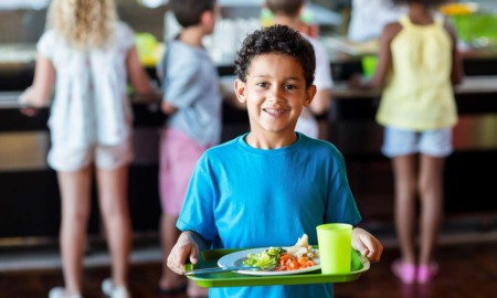 School meals report: happy child holding a tray of school food with other students lined up in background to get food