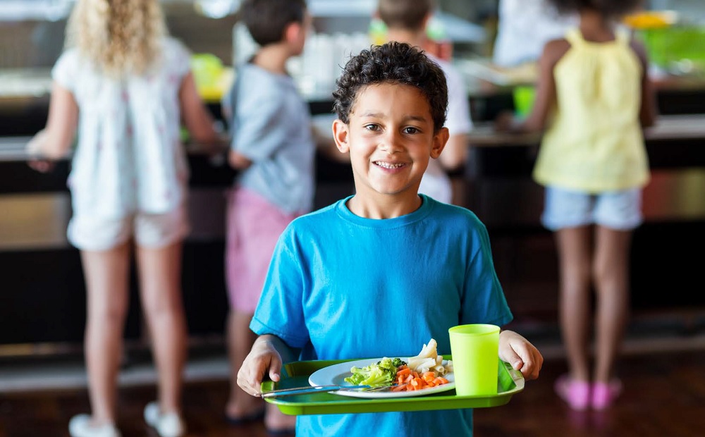 School meals report: happy child holding a tray of school food with other students lined up in background to get food
